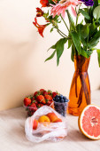 květinové a ovocné složení s kyticí ve váze, bobulemi, grapefruitem a meruňky na dřevěném povrchu na béžovém pozadí
