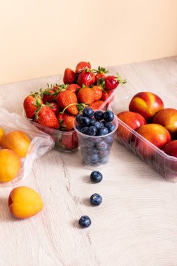 Tahta yüzeydeki plastik kaplarda yaban mersini, çilek, nektarin ve şeftali içeren meyve bileşimi bej rengi.