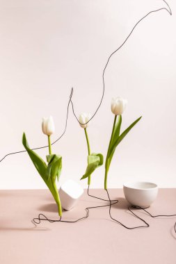 creative floral composition with tulips on wires, cup and square cube isolated on beige clipart