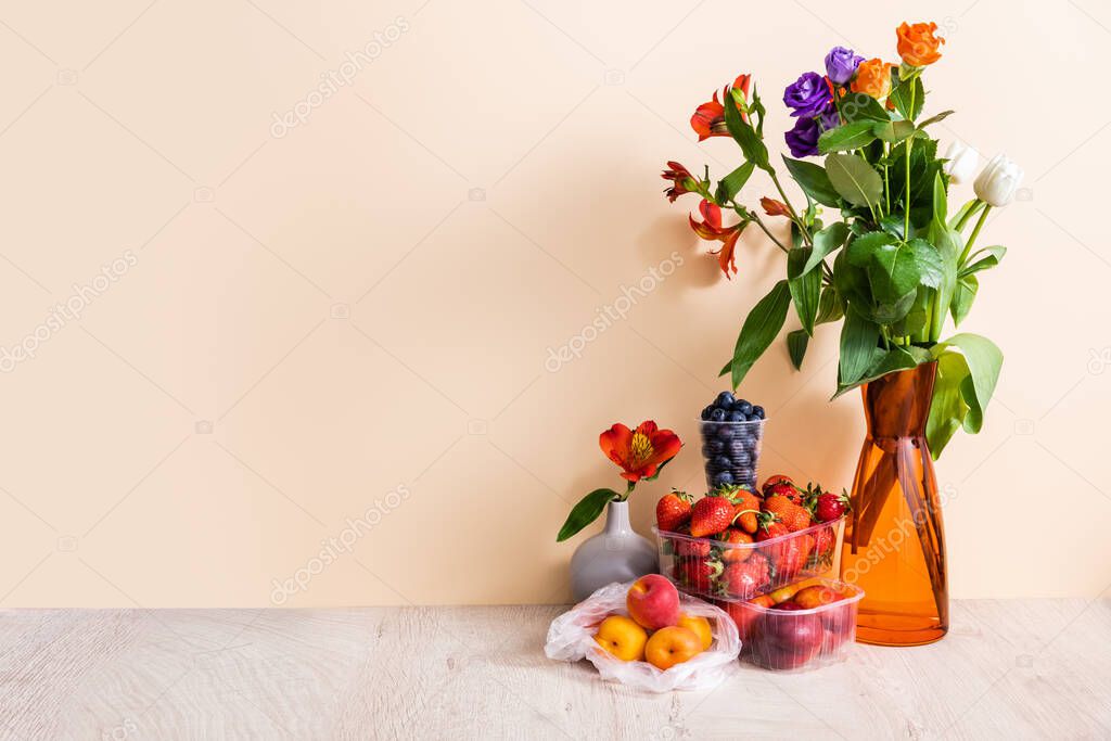 floral and fruit composition with bouquet in vase and summer fruits on wooden surface on beige background
