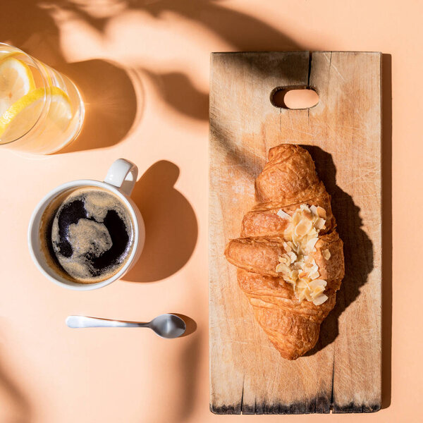 top view of coffee, water and croissant on wooden cutting board for breakfast on beige table