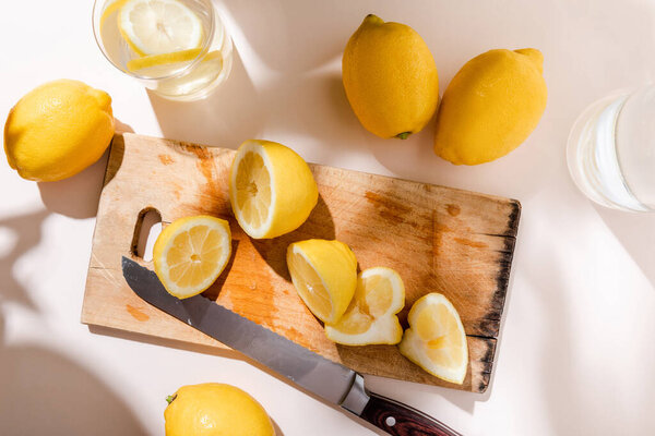 вид сверху на целые и вырезанные лимоны на деревянной доске с ножом и стаканами воды на сером столе
