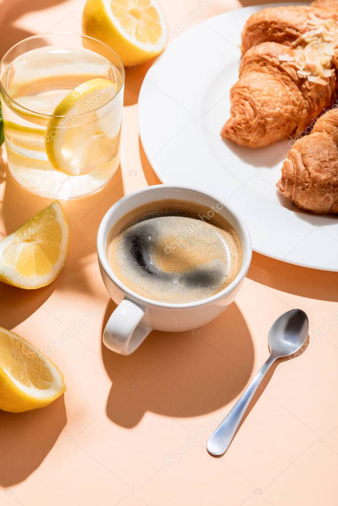 coffee cup with teaspoon, croissants and glass of water with lemon for breakfast on beige table 