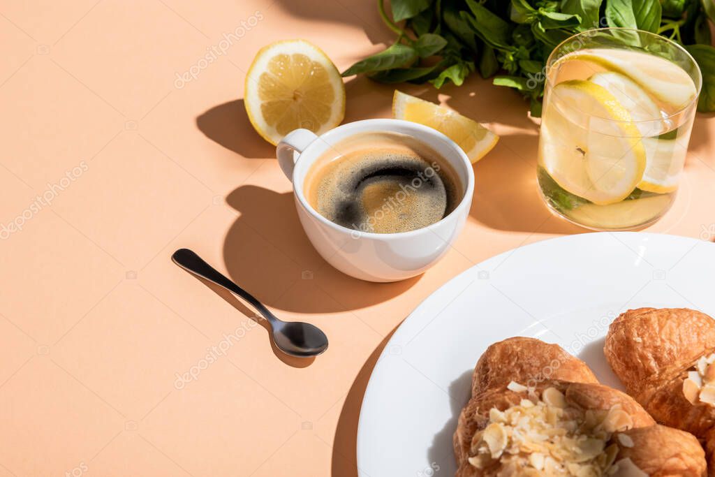coffee, croissants and glass of water with lemon and basil for breakfast on beige table