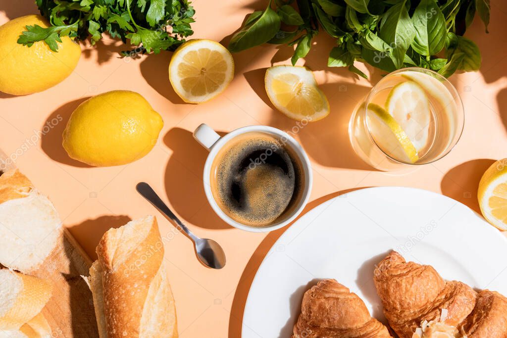 top view of coffee, lemons, greenery, baguette and croissants for breakfast on beige table