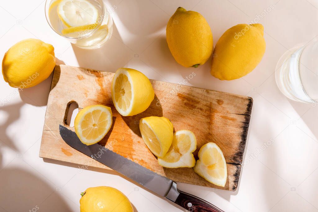 top view of whole and cutted lemons on wooden board with knife and glasses of water on grey table