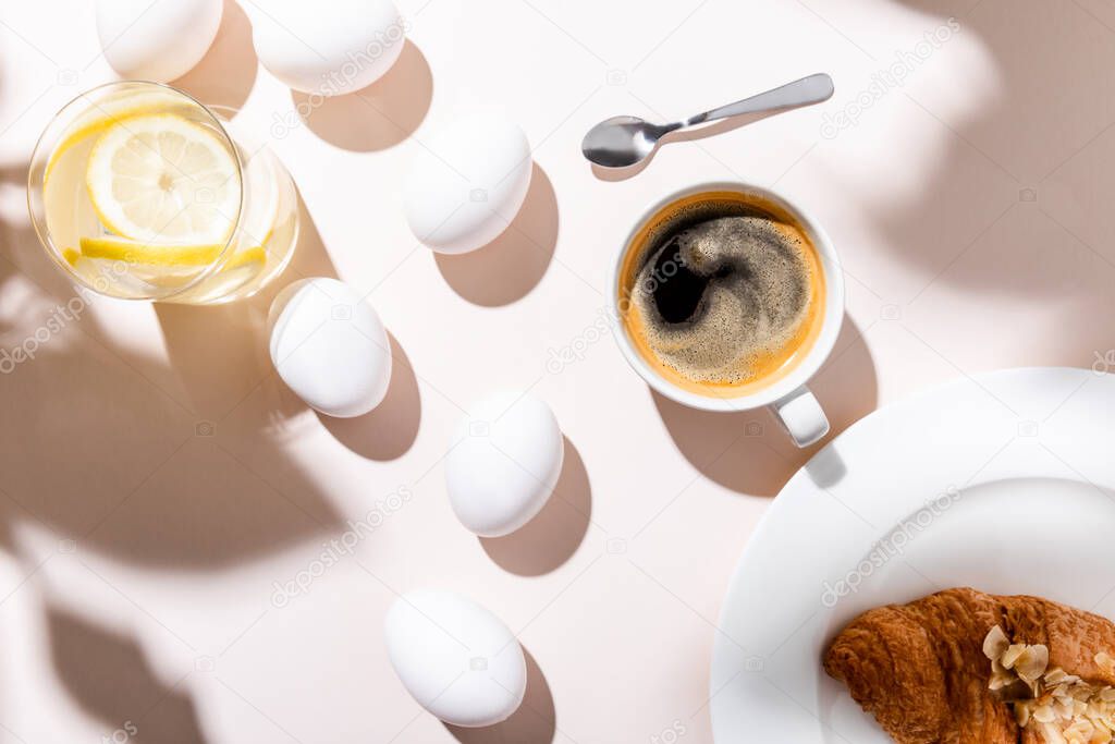 top view of chicken eggs, water with lemon, coffee cup and croissant for breakfast on grey table with shadows