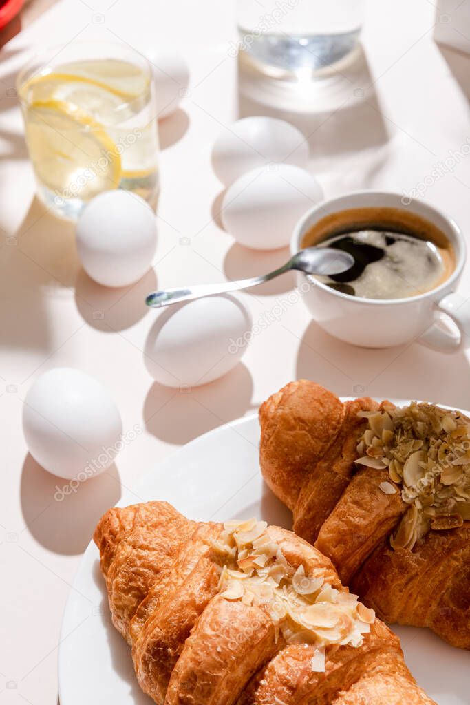 Boiled eggs, two croissants, cup of coffee and glass of water with lemon for breakfast on grey table with shadows 