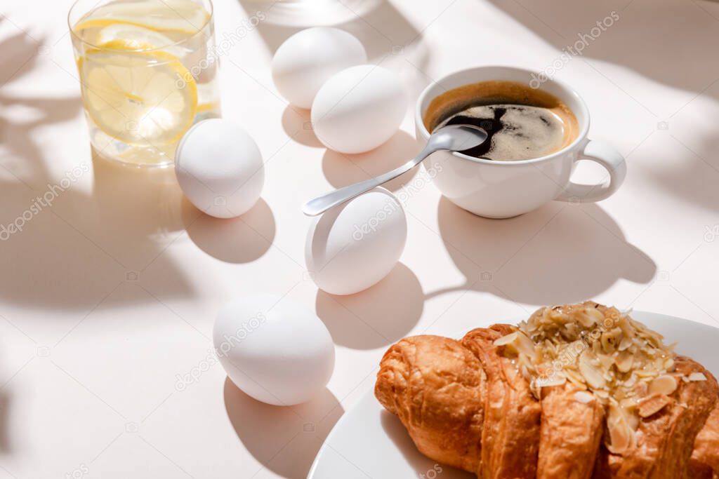 chicken eggs, croissant, cup of coffee and glass of water with lemon for breakfast on grey table with shadows