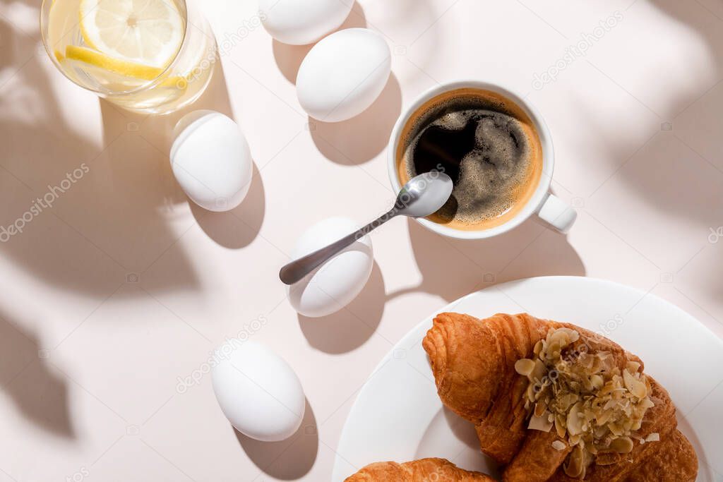 top view of chicken eggs, water with lemon, coffee and croissants for breakfast on grey table with shadows