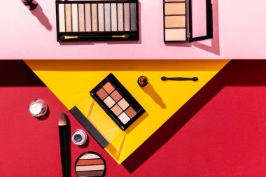 top view of eye shadow palettes and cosmetic brushes near lipstick on crimson, pink and yellow clipart