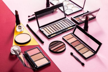 eye shadow palettes and cosmetic brushes near lipsticks and face powder on crimson, pink and yellow clipart