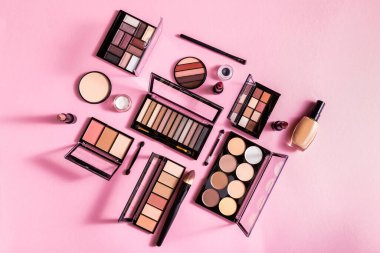 top view of eye shadow and blush palettes near face powder, cosmetic brushes, lipsticks and face foundation on pink clipart