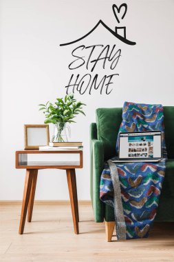 KYIV, UKRAINE - APRIL 14, 2020: stay home lettering near green sofa, blanket, laptop with amazon website and wooden coffee table with green plant clipart