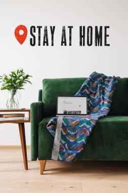 KYIV, UKRAINE - APRIL 14, 2020: green sofa, blanket and laptop with airbnb website near wooden coffee table with green plant and stay at home lettering  clipart