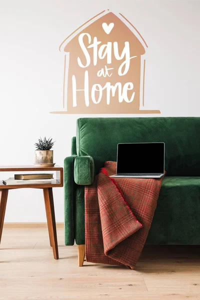 green sofa with blanket and laptop near wooden coffee table and stay at home lettering