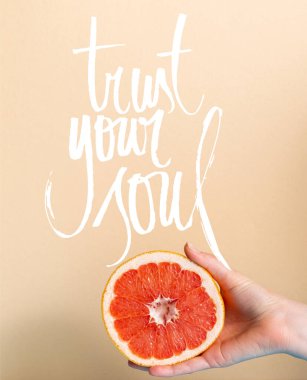 cropped view of female hand with juicy grapefruit half near trust your soul lettering on beige  clipart