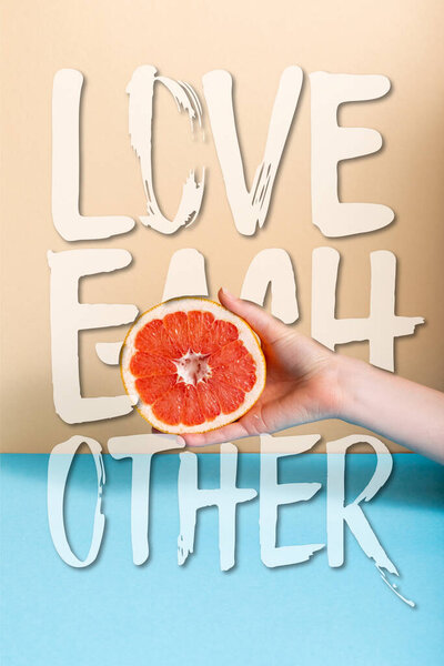 cropped view of female hand with juicy grapefruit half near love each other lettering on beige and blue 