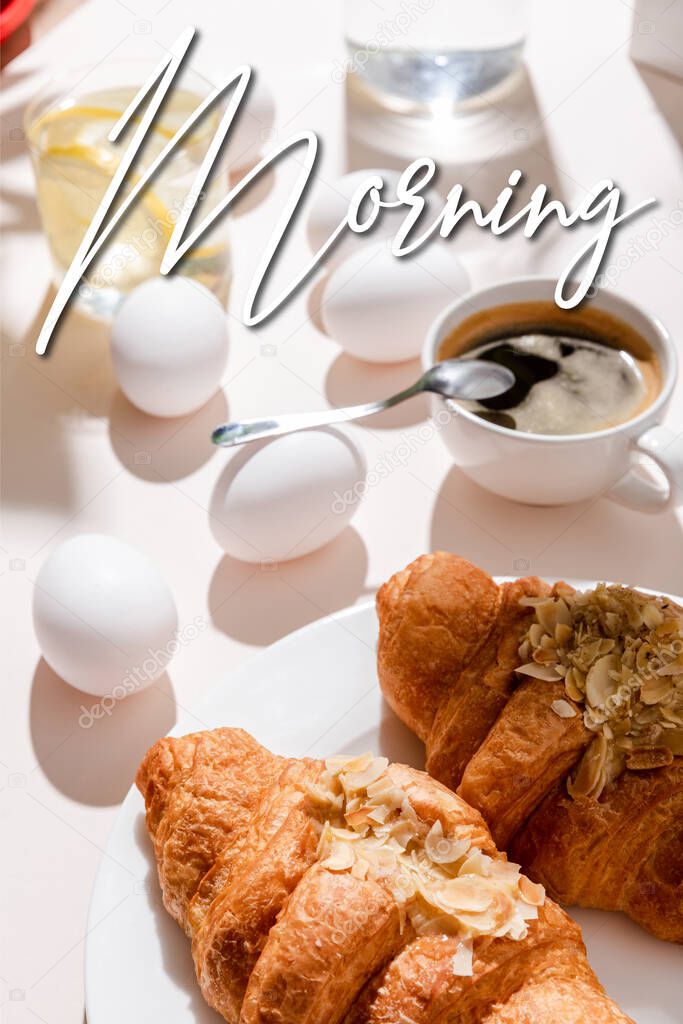Boiled eggs, two croissants, cup of coffee and glass of water with lemon for breakfast on grey table with morning lettering 