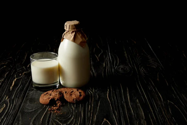 Schokoladenkekse, frische Milch in Glas und Flasche mit Papier auf schwarzem Hintergrund umwickelt — Stockfoto