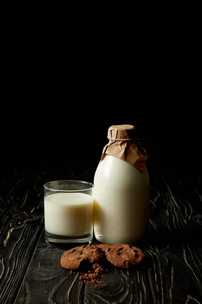 Frischmilch im Glas, mit Papier umwickelte Milchflasche und Schokoladenkekse auf schwarzem Hintergrund — Stockfoto