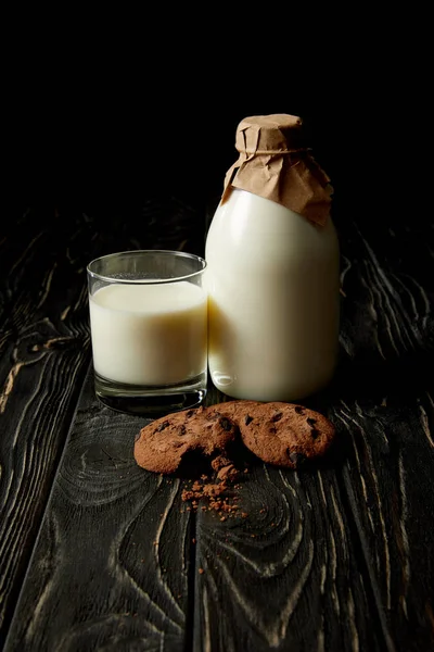 Vista de cerca de galletas de chocolate, leche fresca en vidrio y botella envuelta por papel sobre fondo negro - foto de stock