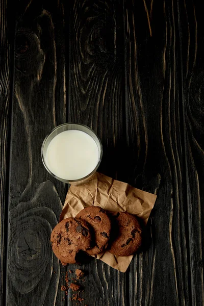 Top view of chocolate cookies on crumpled paper and milk glass on black wooden surface — Stock Photo