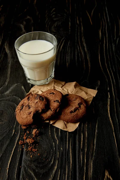 Vista de cerca de galletas de chocolate en papel arrugado y vaso de leche en la superficie de madera negra - foto de stock