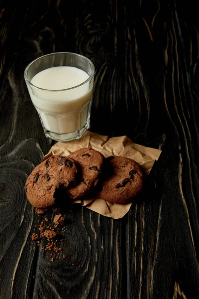 Vista close-up de biscoitos de chocolate em papel amassado e vidro de leite na superfície de madeira preta — Fotografia de Stock