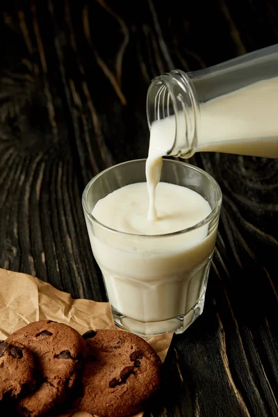 Молоко, наливаемое в стакан из бутылки и шоколадное печенье на мятой бумаге — стоковое фото