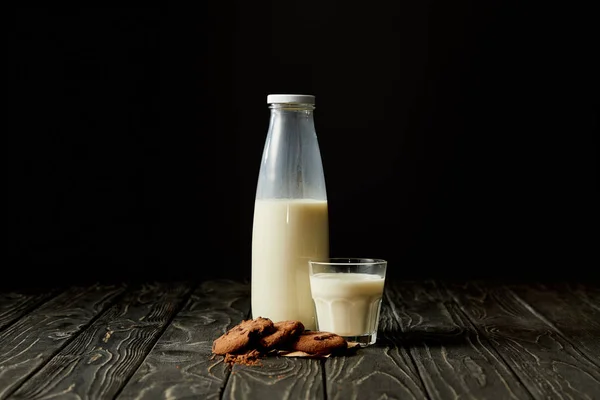 Galletas de chocolate, leche en botella y vidrio sobre fondo negro - foto de stock