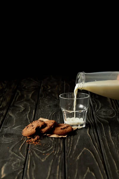 Imagen recortada de la persona que vierte leche en el vaso de galletas de botella y chocolate en la superficie de madera negro - foto de stock