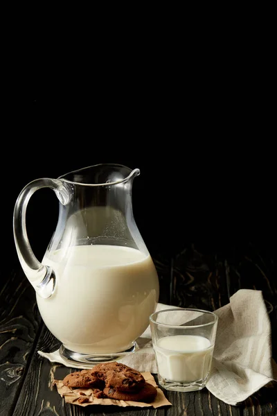 Leche fresca en jarra y vaso, galletas de chocolate y saco sobre fondo negro - foto de stock