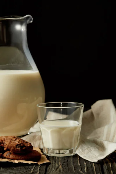 Vista parziale del latte fresco in brocca e vetro, biscotti al cioccolato e sacco su fondo nero — Foto stock