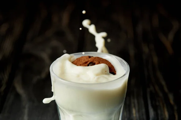 Vue rapprochée de biscuit au chocolat en verre de lait avec éclaboussures sur la surface en bois noir — Photo de stock