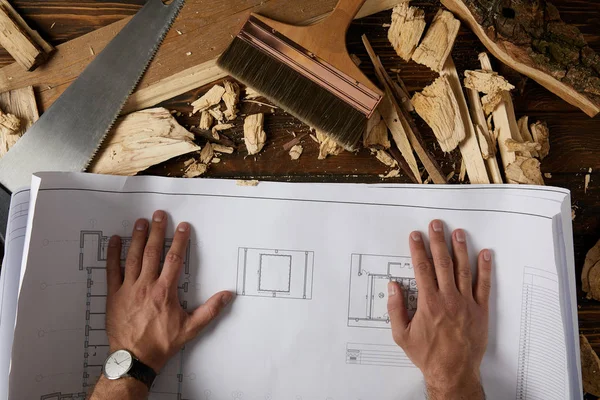 Cropped shot of architect hands on blueprint at table with paintbrush, handsaw and wooden chips on table — Stock Photo
