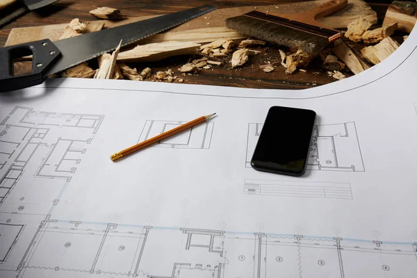 Closeup view of smartphone and pencil on blueprint, handsaw and paintbrush with wooden pieces on table — Stock Photo