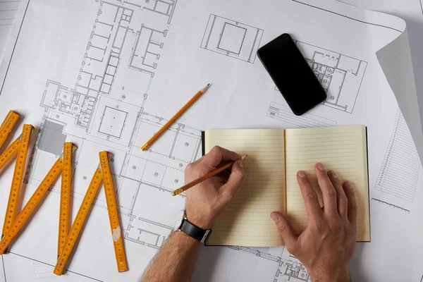Imagen recortada de arquitecto escribiendo en libro de texto en blanco sobre plano con lápiz, smartphone y medidor plegable - foto de stock