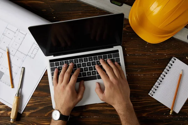 Cropped image of architect typing on laptop at table with chisel, spirit level, protective helmet, blueprint, textbook and pencils — Stock Photo