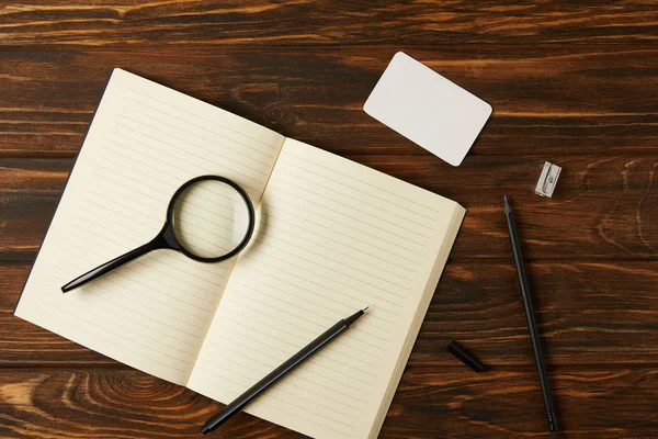 Top view of magnifying glass, blank notebook and office supplies on wooden table — Stock Photo