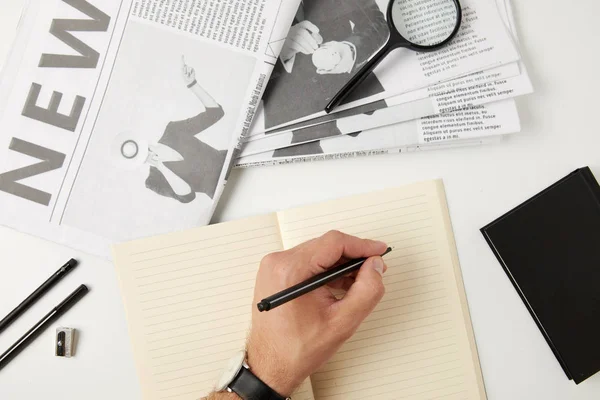Partial top view of person writing in blank notebook, newspapers, magnifying glass, notebook and office supplies on grey — Stock Photo