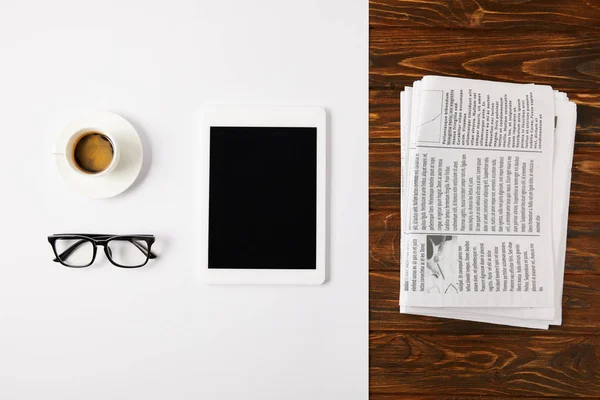 Top view of eyeglasses, coffee cup, and digital tablet with blank screen on white and pile of newspapers on wooden background — Stock Photo