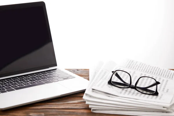 Gafas graduadas en periódicos y laptop con pantalla en blanco sobre superficie de madera, sobre blanco - foto de stock