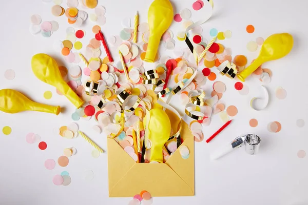 Vista dall'alto di pezzi di coriandoli, palloncini e busta gialla su superficie bianca — Foto stock