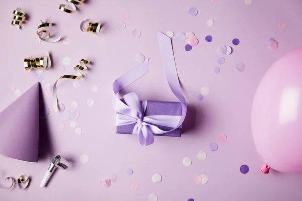 Top view of one violet present box, balloon, party hat and confetti pieces on surface — Stock Photo