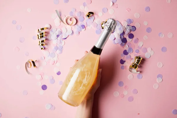 Cropped image of woman holding bottle of champagne above confetti on surface — Stock Photo