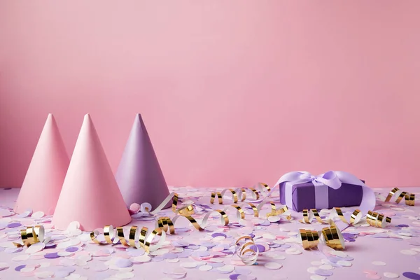 Party hats, gift box and confetti pieces on violet tabletop — Stock Photo