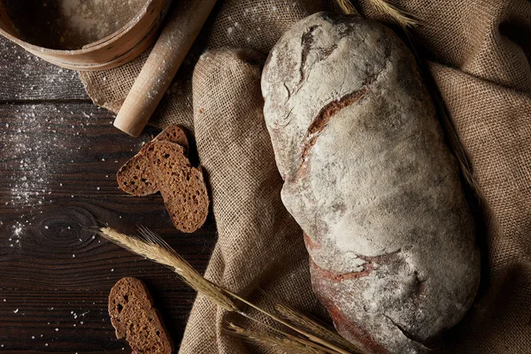 Vista sopraelevata di pane, frumento, mattarello, setaccio e sacco su tavola di legno ricoperta da farina — Foto stock