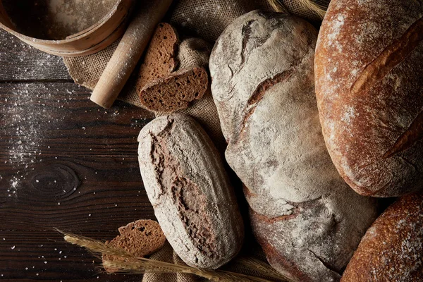 Top view of various types of bread, wheat, rolling pin, sieve and sackcloth on wooden table covering by flour — Stock Photo