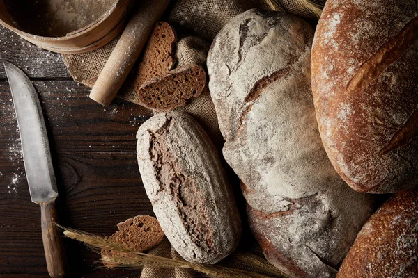 Vista elevada de varios tipos de pan, trigo, rodillo, tamiz y saco en la mesa de madera - foto de stock